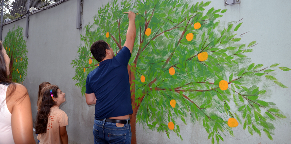 A man paints a mural of an orange tree on a wall, while children watch.