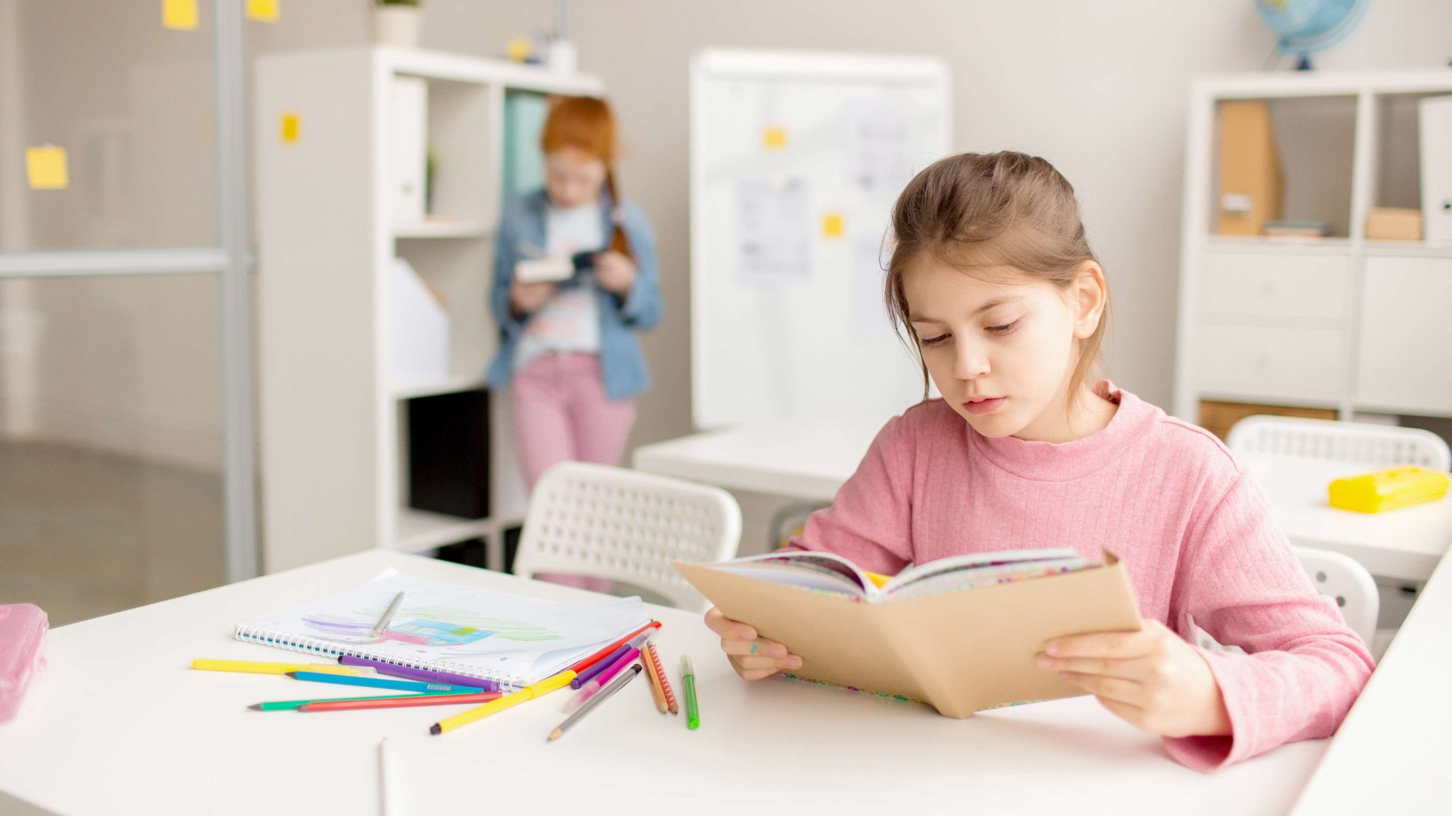 Preschool student attentively reading a book at her desk, surrounded by colorful art supplies.
