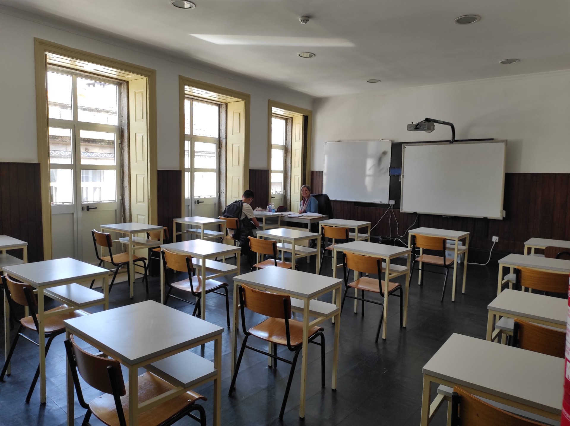 Empty classroom with tables and chairs, ready for students. Whiteboard and projector are visible.