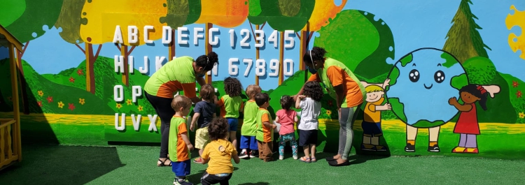 Preschool children learning the alphabet and numbers with their teachers in front of a colorful mural.