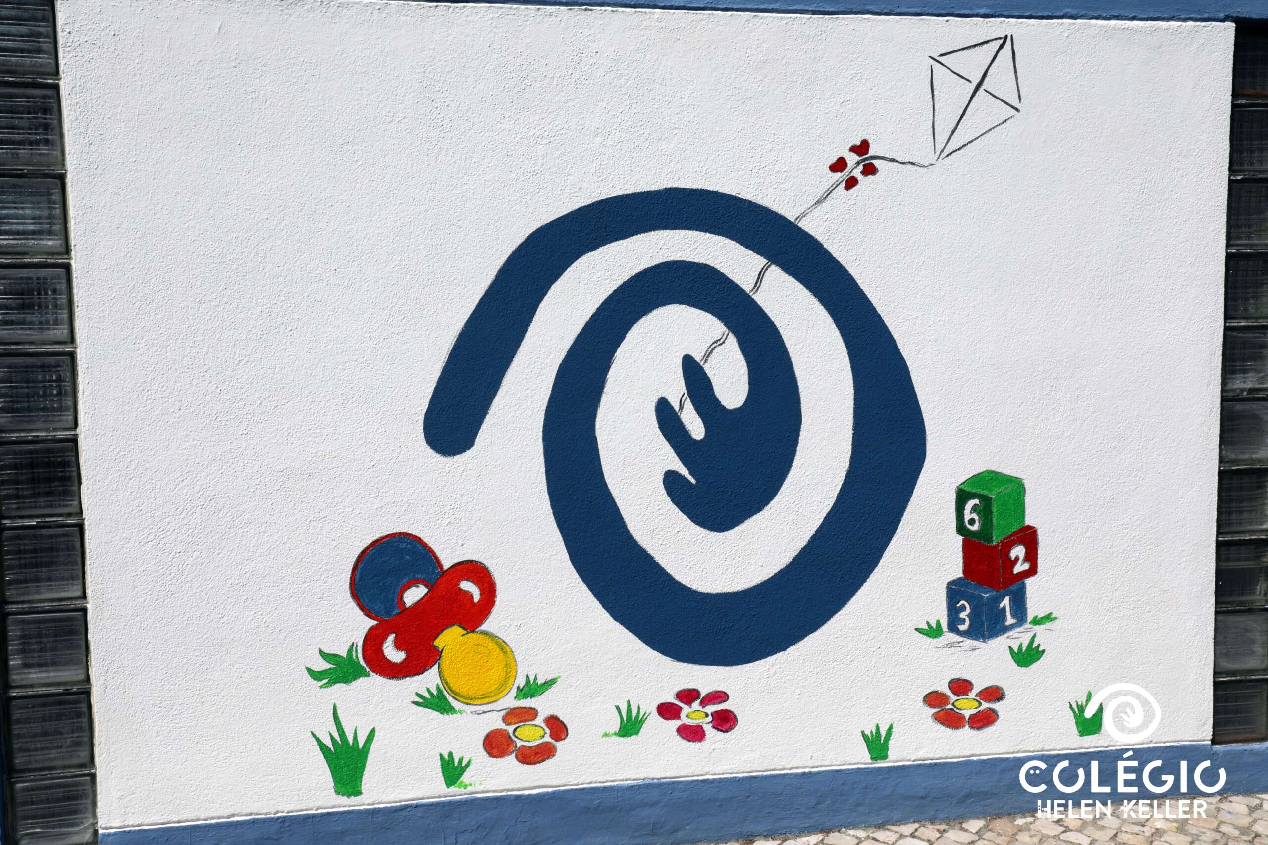 Colorful mural on the exterior wall of Colégio Helen Keller preschool, featuring playful imagery such as a spiral, kite, building blocks, and pacifier.