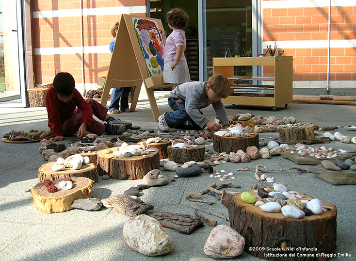 Preschool children engaged in a nature-based activity, sorting and arranging rocks and shells on tree stumps.