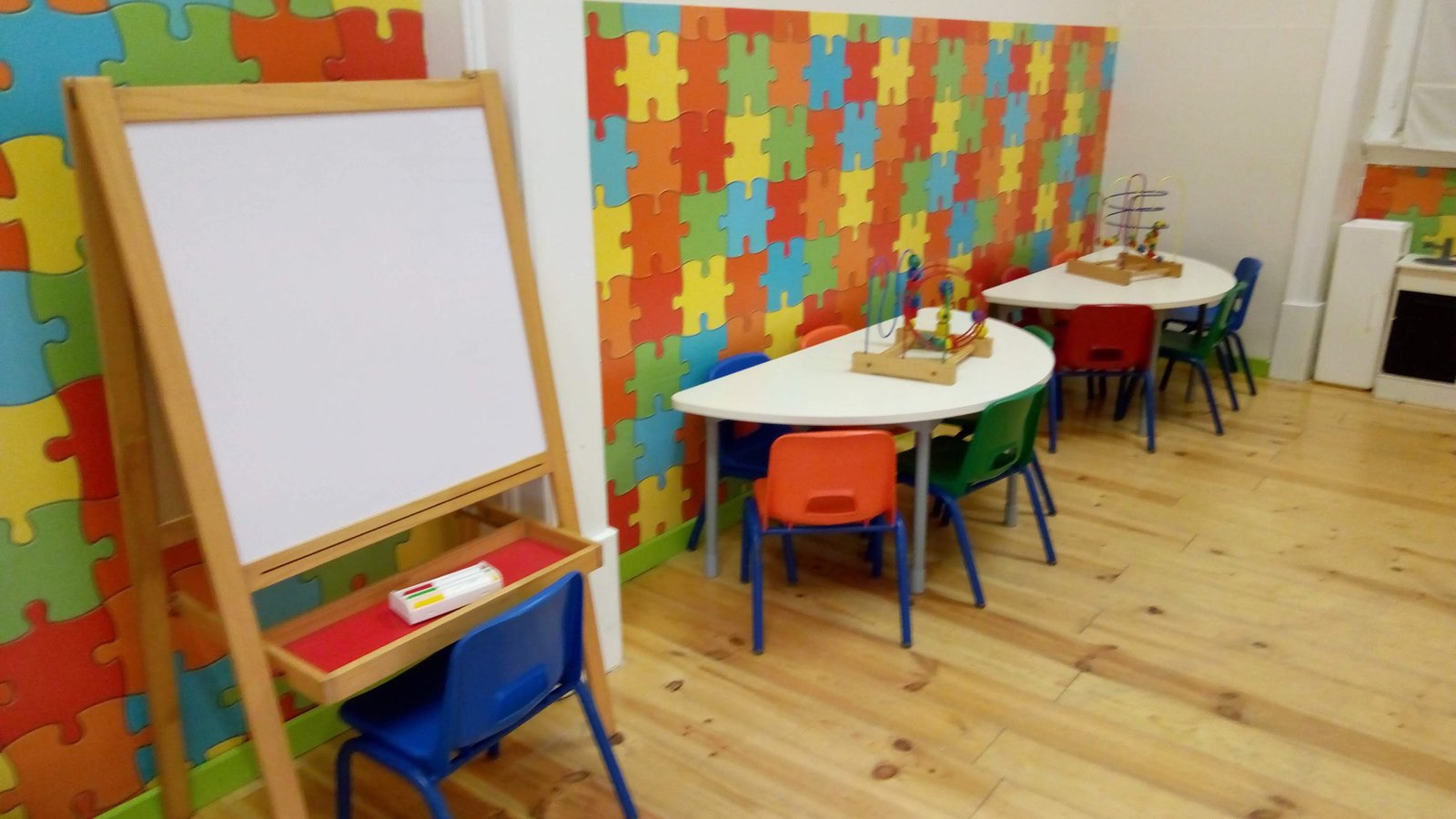 Preschool classroom with colorful puzzle wall, small tables, chairs, and a whiteboard easel.