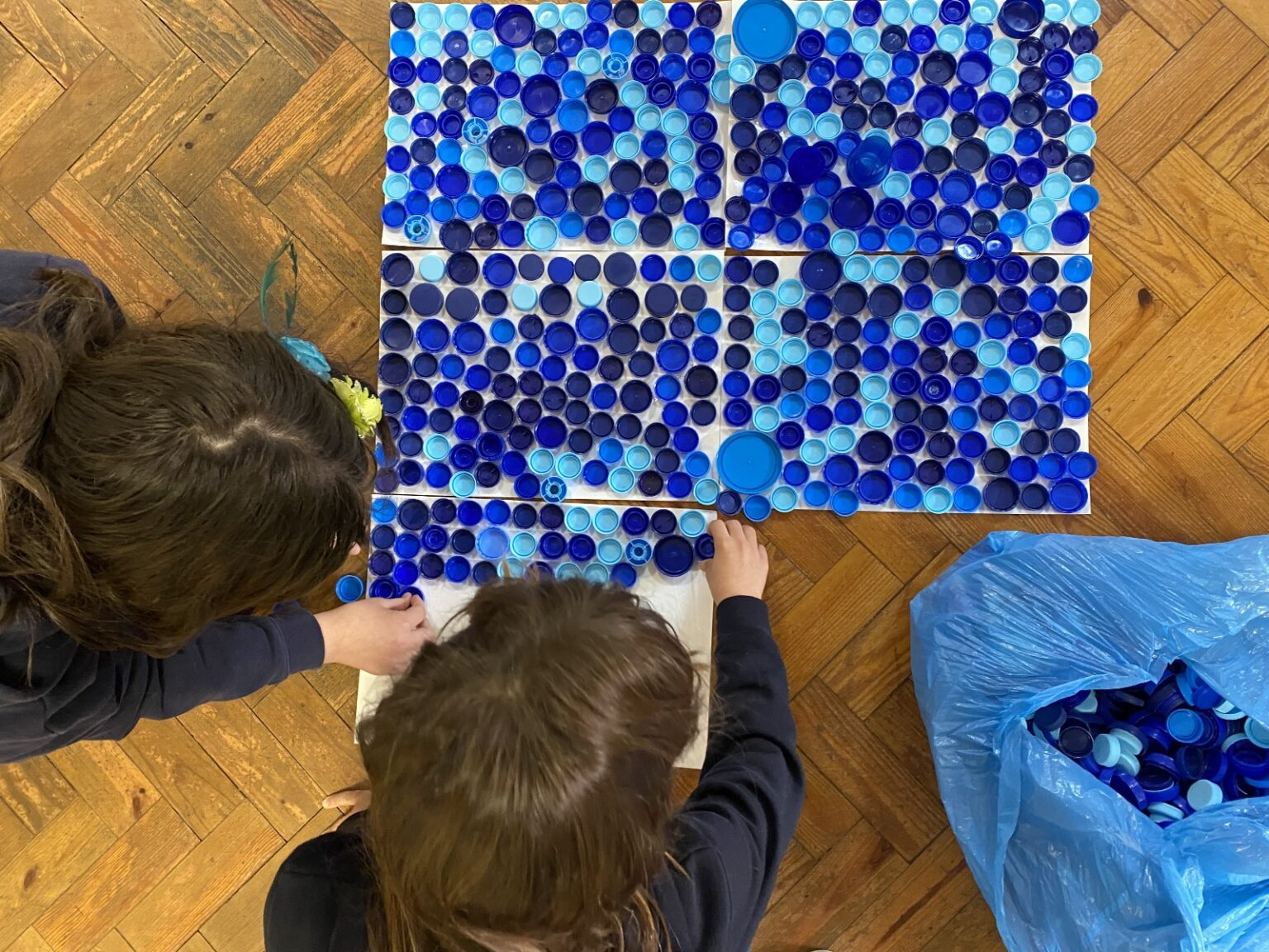 Two children collaborate on an art project using blue bottle caps, creating a colorful mosaic on a wooden floor.