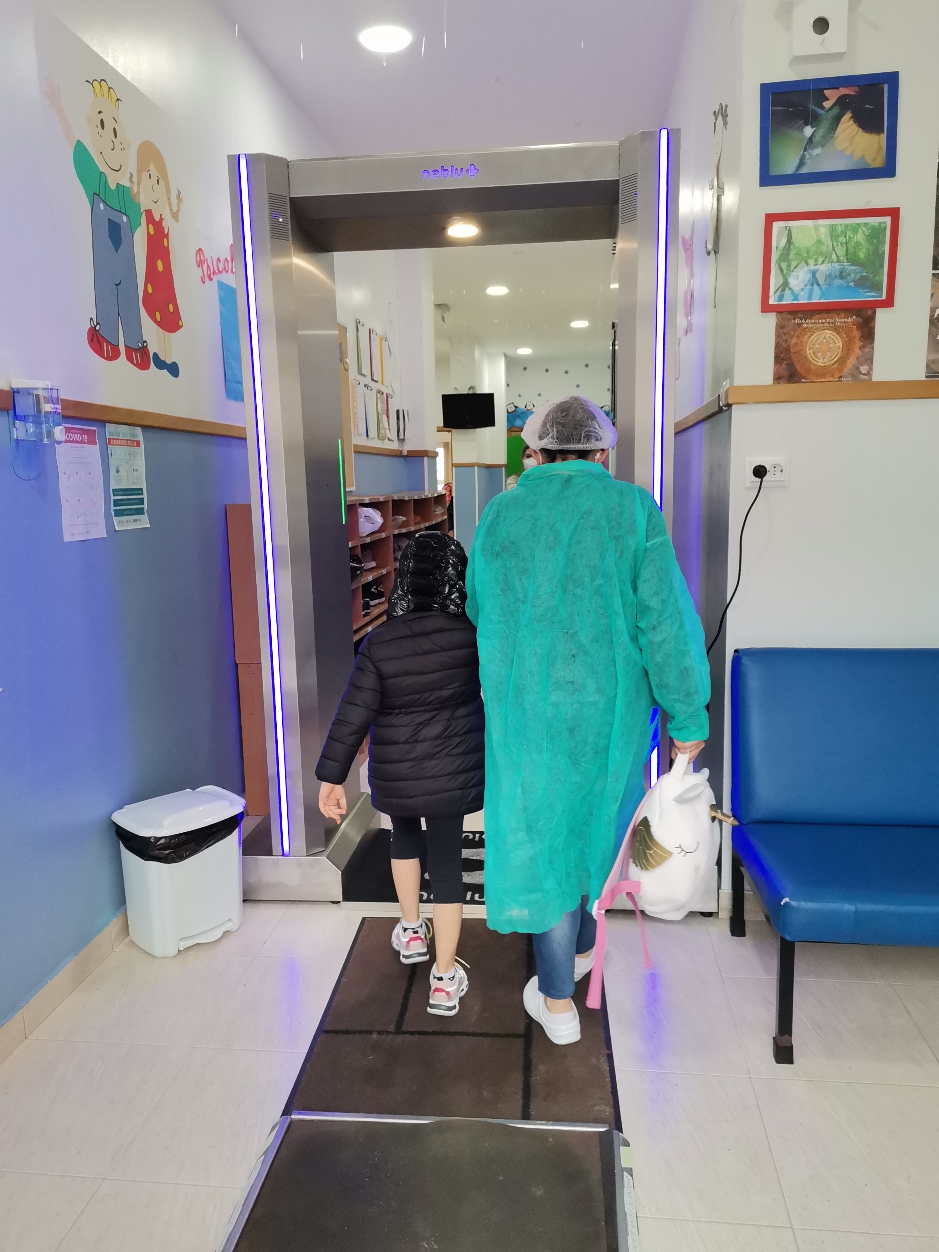 Child and staff member walking through a sanitizing entrance at Creche E Jardim De Infância Cosminho E Damiãozinho Lda preschool.