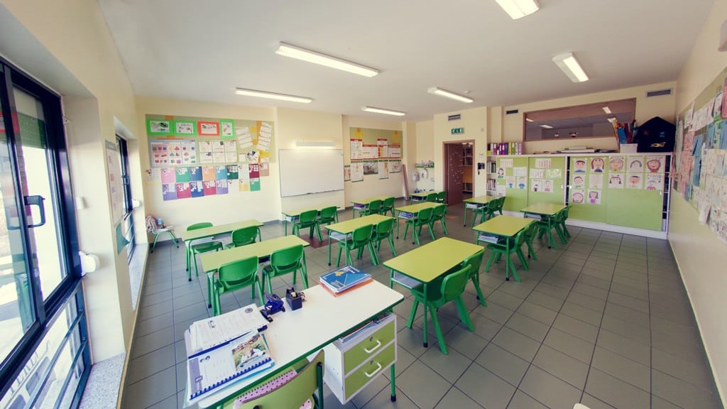 Preschool classroom with green tables and chairs, educational materials on the walls, and a whiteboard.