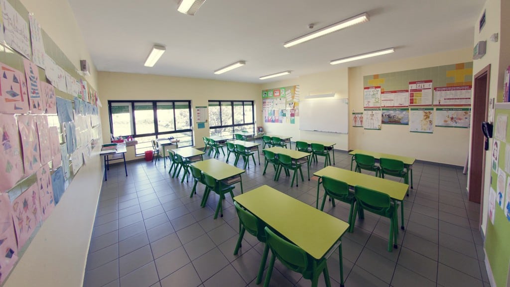 Preschool classroom with green tables and chairs, educational posters, and children's artwork.