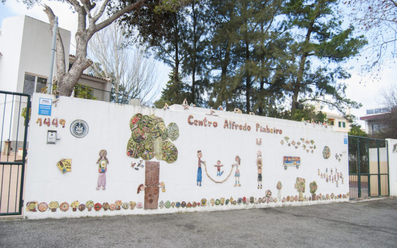 Colorful tile mural depicting children playing and a school bus, located on the exterior wall of Centro Alfredo Pinheiro preschool.