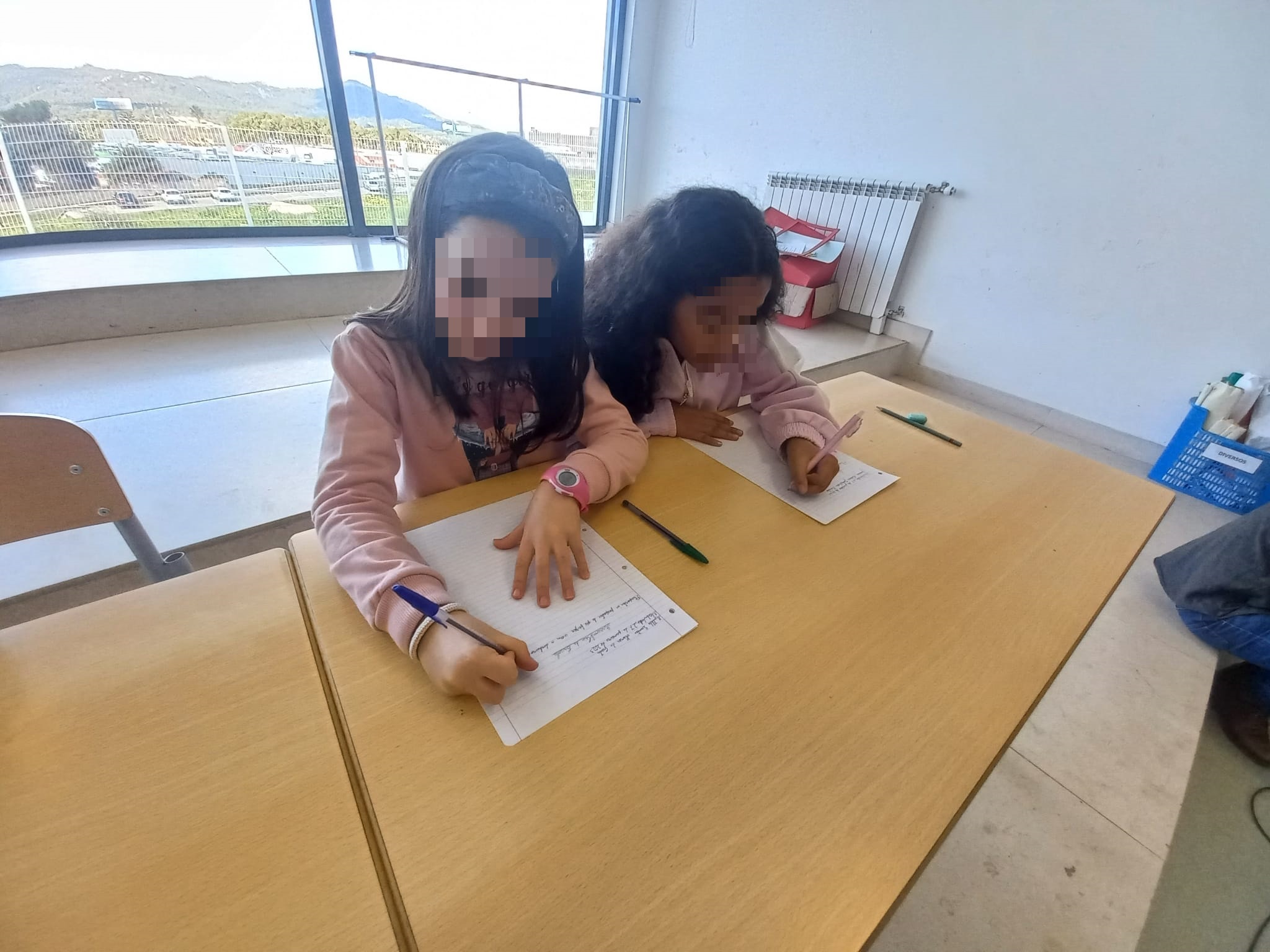 Two students are sitting at desks in a classroom, writing in notebooks.