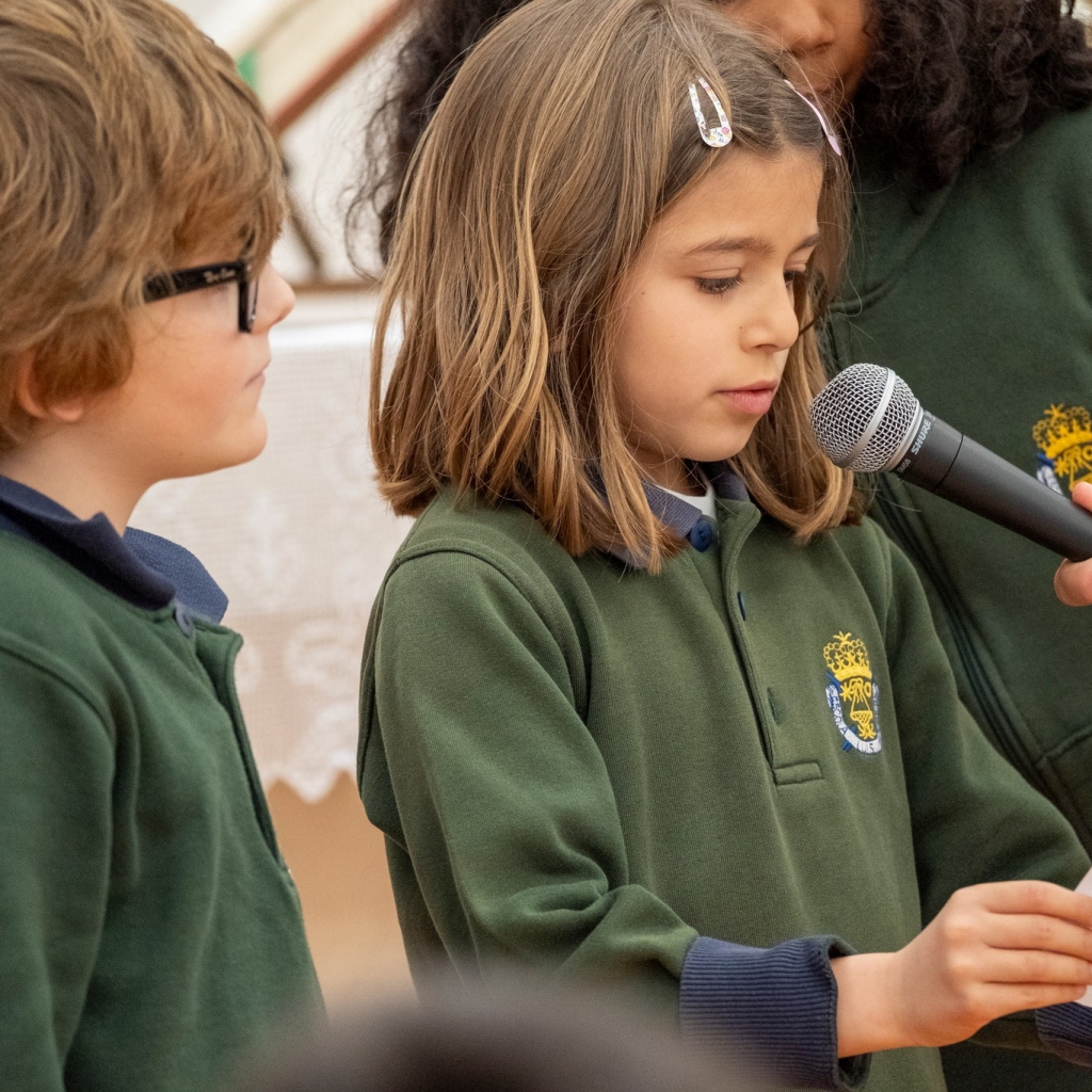 Two students in green school uniforms participate in a school event, one speaking into a microphone.