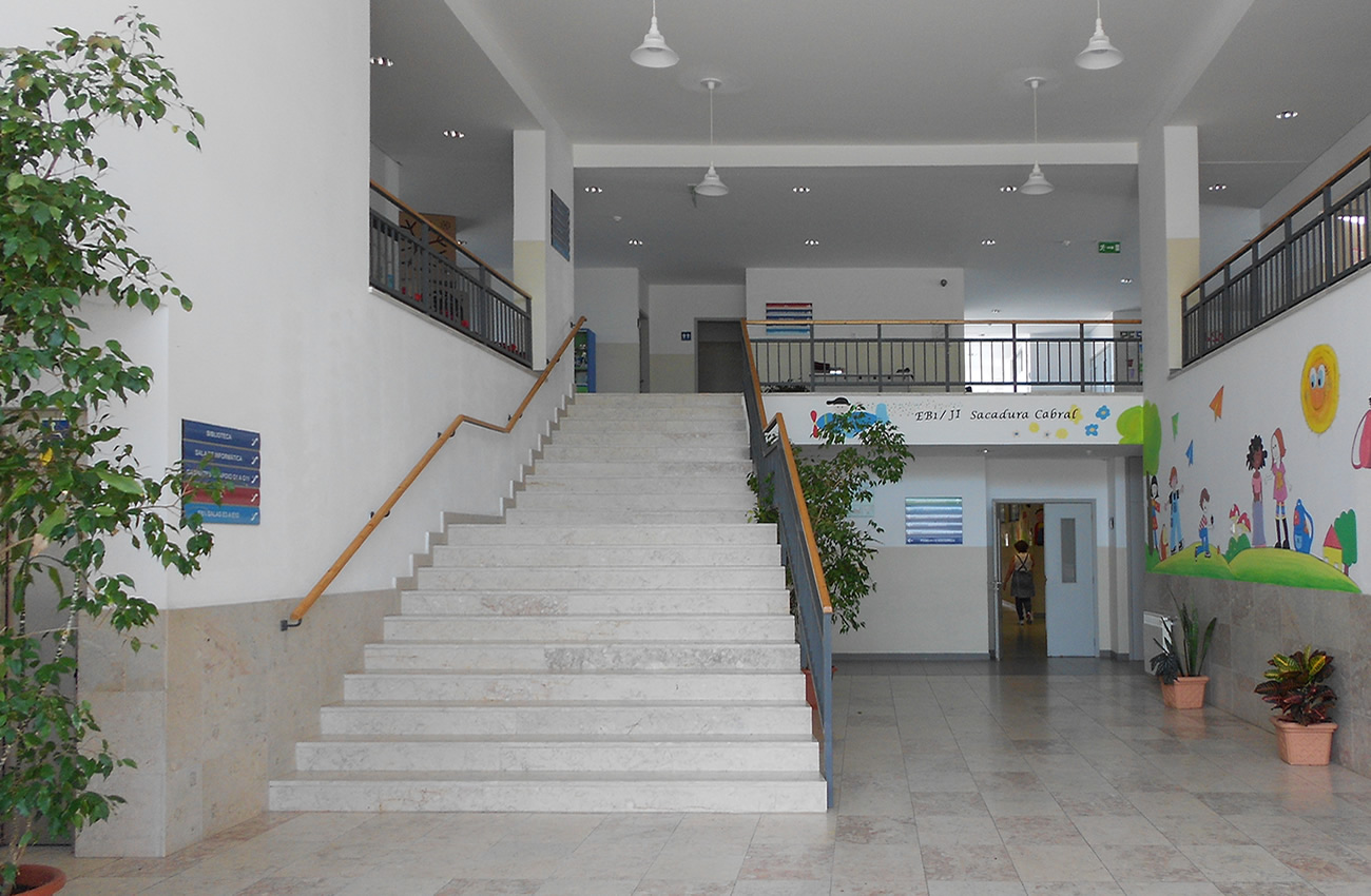 Interior view of JI 2 Brandoa preschool, showing a staircase with light-colored stone steps and wooden handrails. There are plants in pots and colorful murals on the walls.