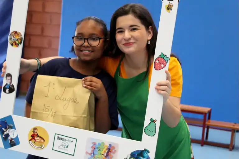 A teacher and student holding a decorated frame. The student is holding a paper bag with the words "1lugar 15/17" written on it.
