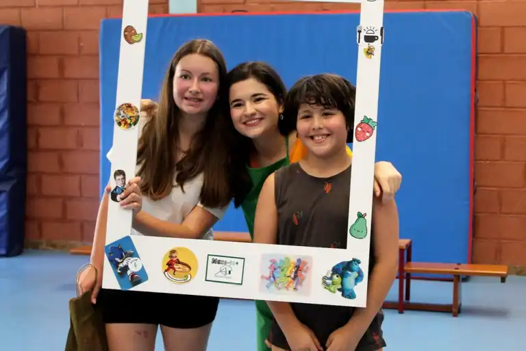 Three children hold a large photo frame with cartoonish food and character stickers. The frame is decorated with drawings of fruits, vegetables, and cartoon characters. The children are smiling and appear to be having fun.