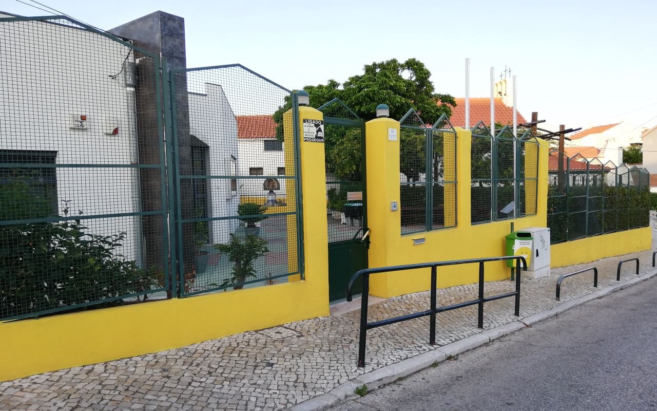 Exterior view of A Nossa Creche preschool, showing a yellow wall, green fence, and gated entrance.