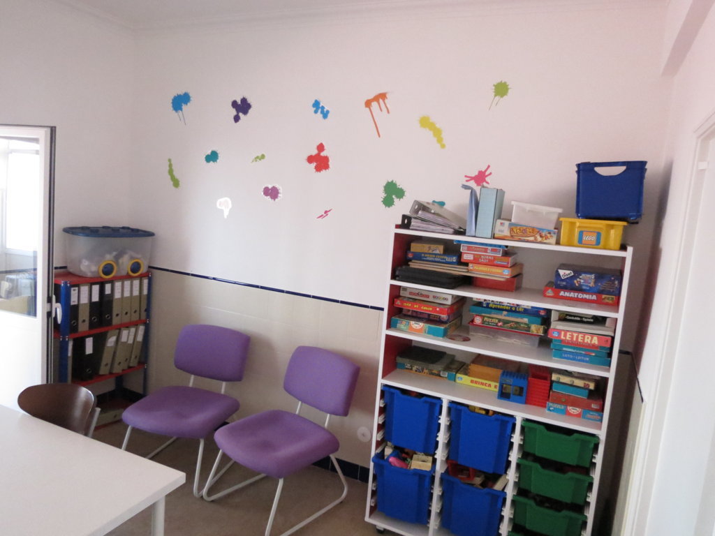 Preschool classroom with colorful paint splatters on the wall, shelves of educational games and toys, and purple chairs.