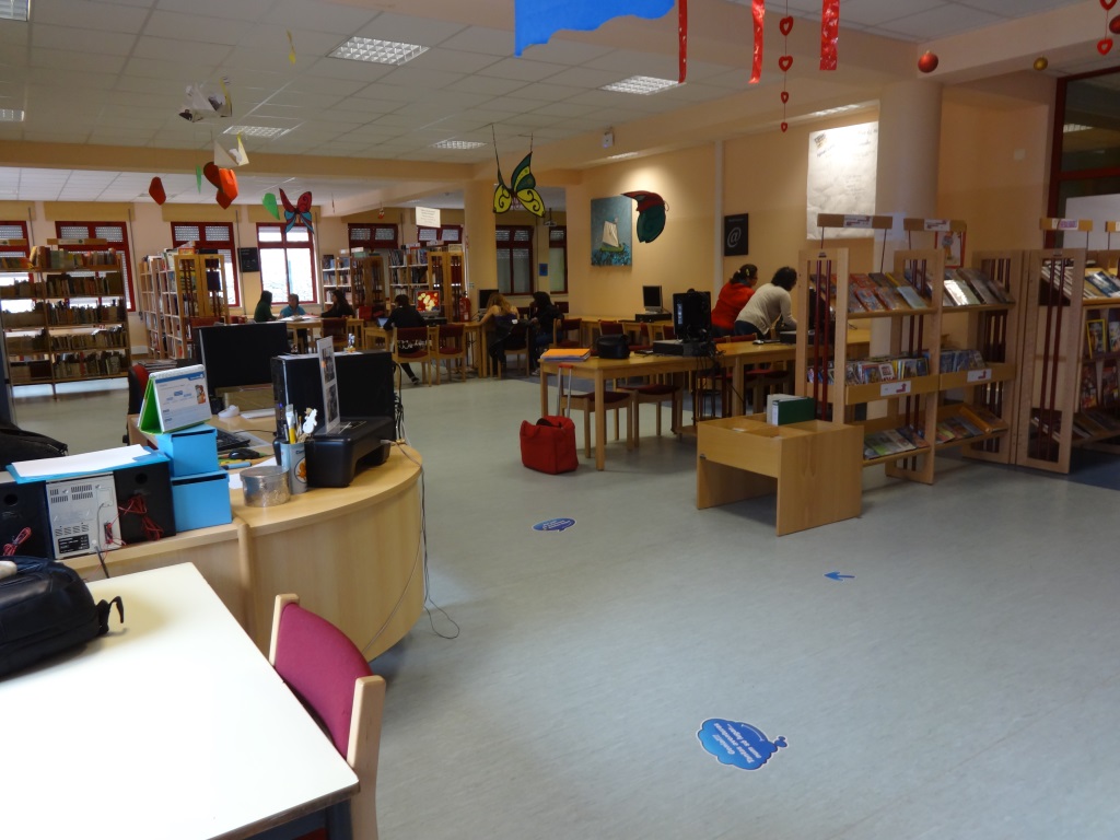 Interior view of the Escola Básica Nun'Álvares library, showing bookshelves, computers, and students. 