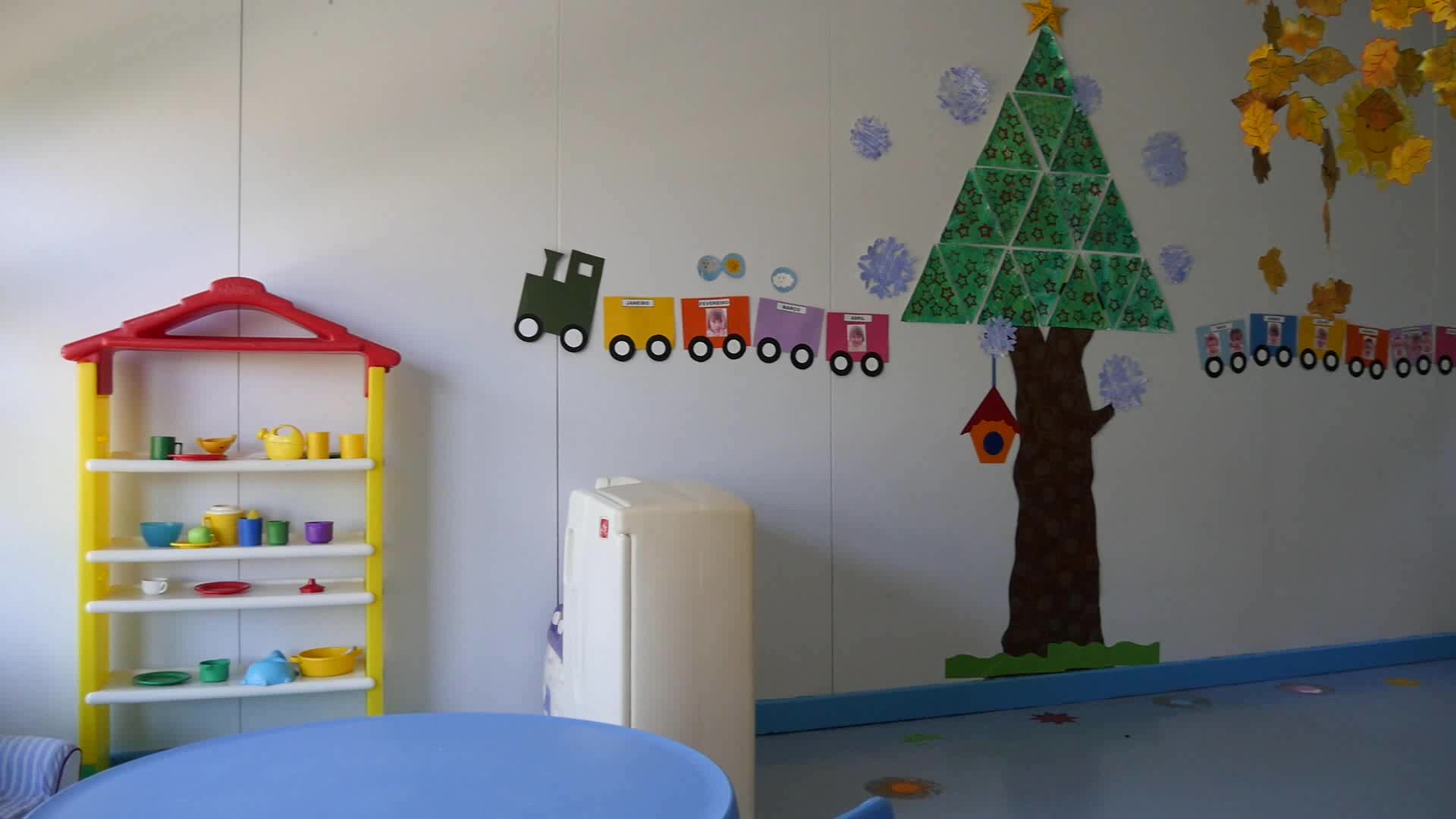 Preschool classroom with a toy shelf, play kitchen, and a wall decorated with a train and Christmas tree theme.