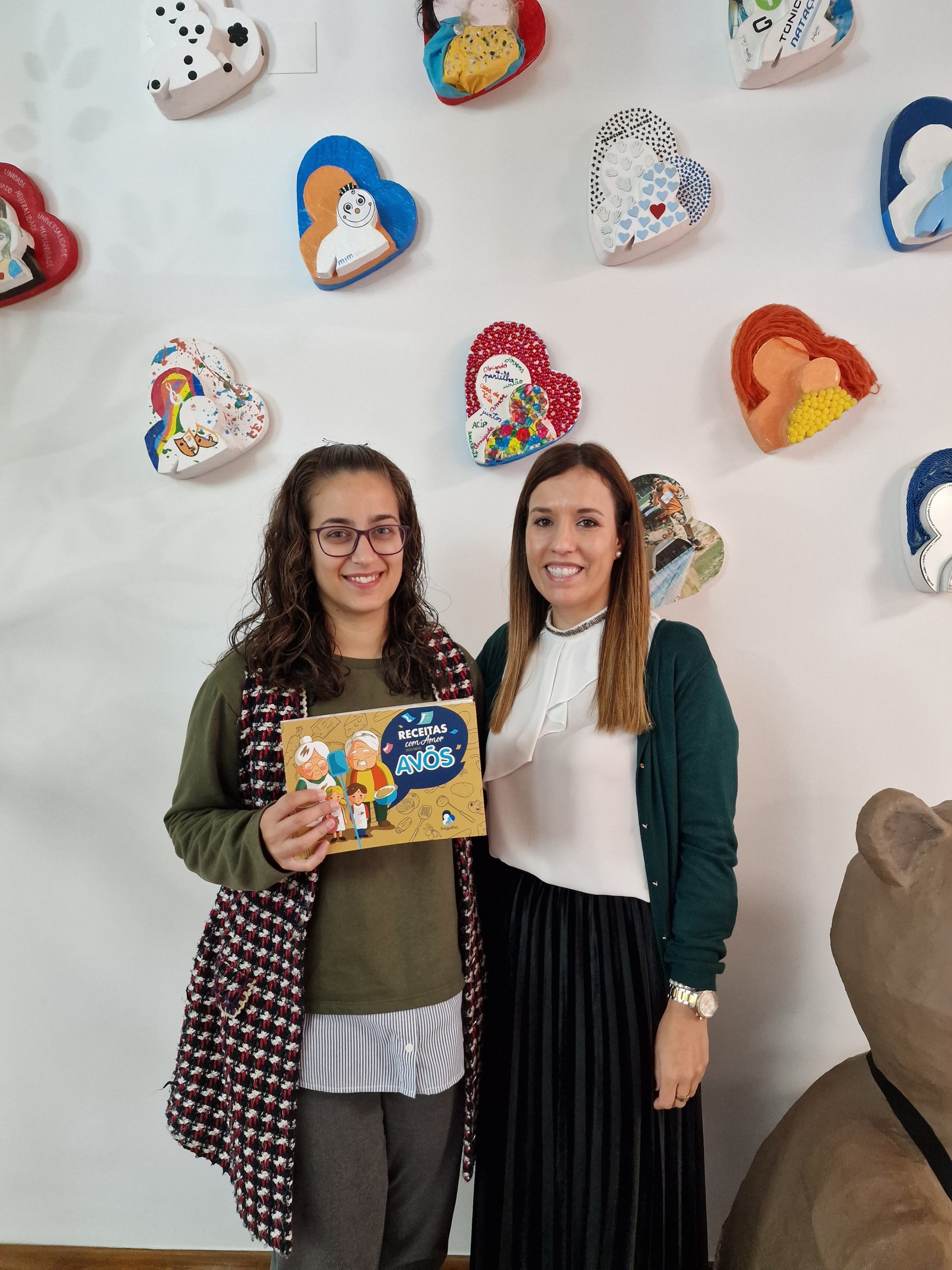 Two women holding a book titled "Recipes with Love for Years" at Bogalha school.