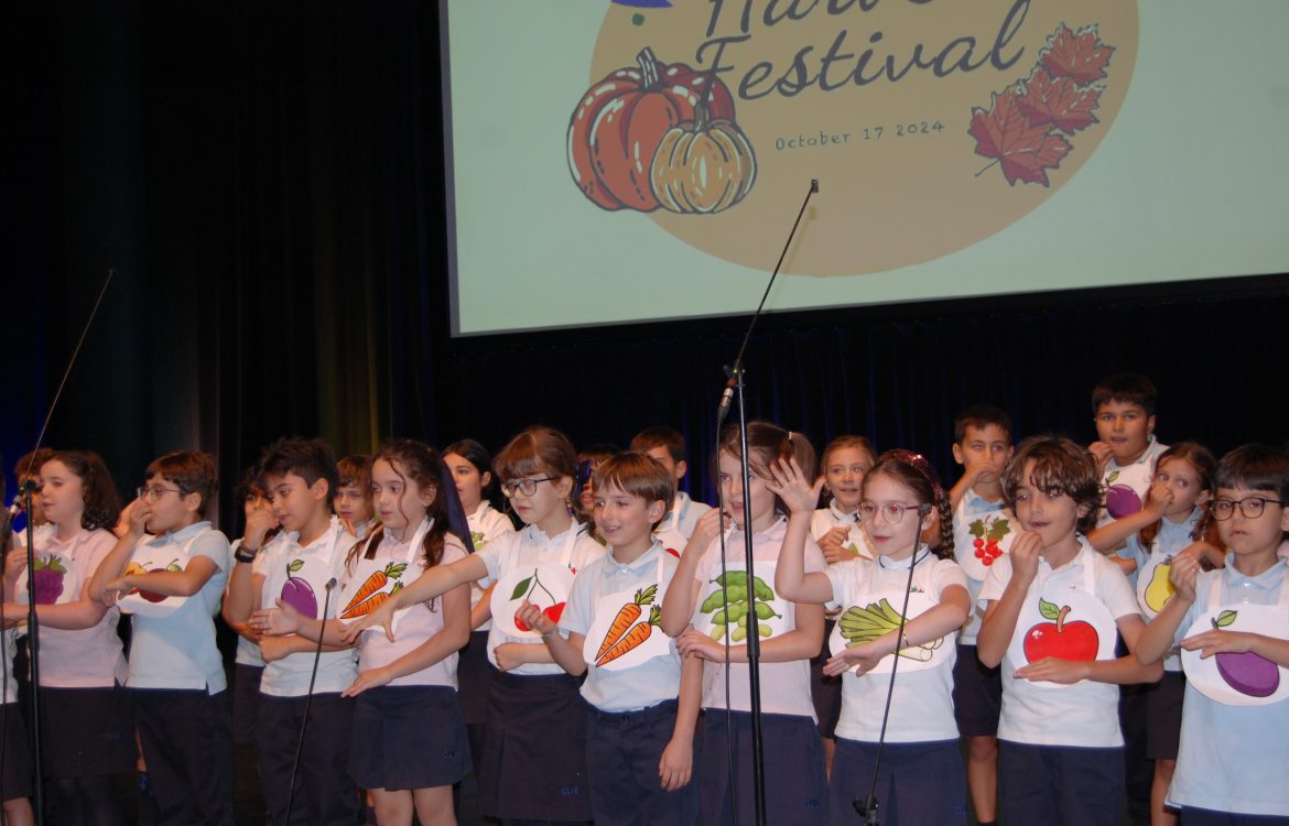 Students in a school Harvest Festival, wearing aprons with fruit and vegetable designs, singing on stage.