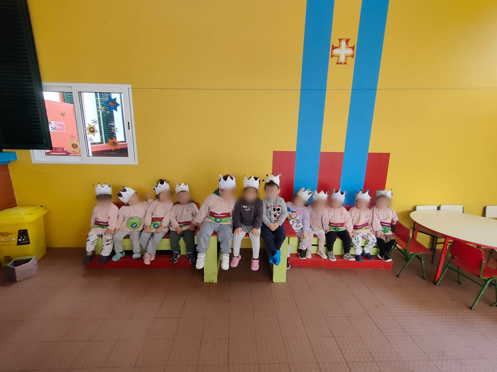 Preschool children wearing crowns sit on a bench in a classroom.