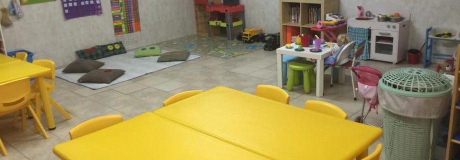 Preschool classroom with yellow tables and chairs, play area with pillows, and toy kitchen