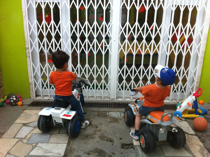 Two preschool children playing on toy cars outside the Colégio O Rezingão.