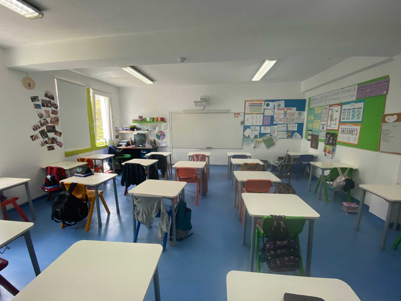 Preschool classroom with colorful desks and chairs, educational materials on the walls, and student backpacks.