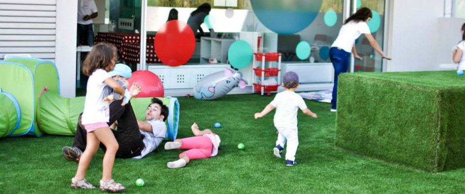 Preschool children playing on a grassy playground with colorful toys and equipment.