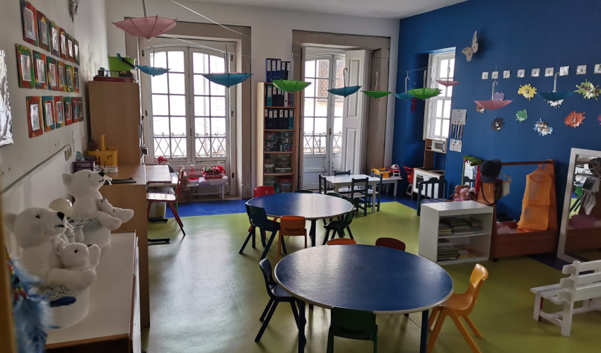 Preschool classroom with colorful tables and chairs, decorated with hanging umbrellas and children's artwork.